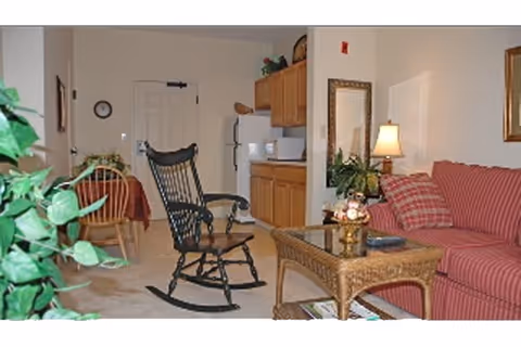 Interior view of a senior living facility room featuring a red striped sofa with plaid pillows, a wicker coffee table with decorative items, a black rocking chair, a small dining table with chairs, and a kitchenette with wooden cabinets and a refrigerator.
