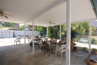 Covered outdoor patio area with multiple chairs and tables arranged under a white roof supported by white pillars. Ceiling fans are mounted on the roof. The patio overlooks a fenced yard with trees, plants, and a shed in the background.