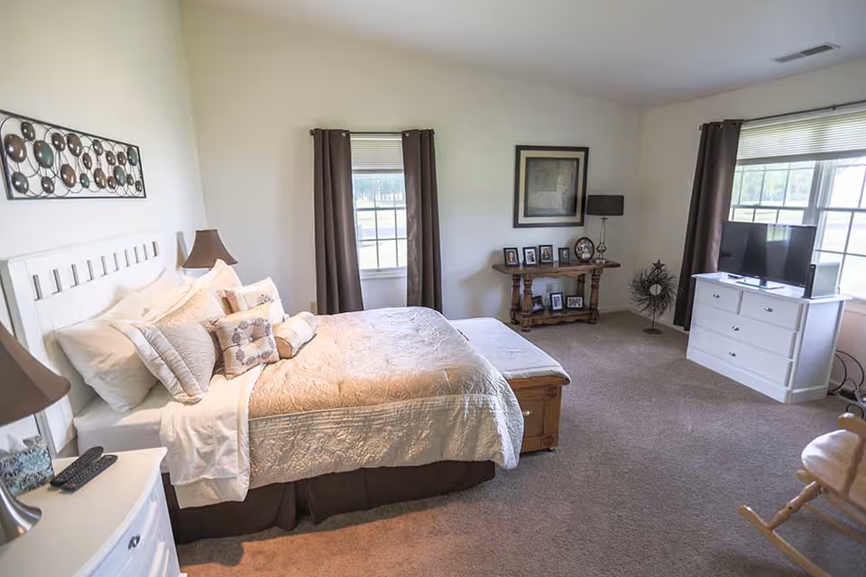 Well-lit furnished bedroom with a large bed, bedside tables, a dresser with TV, windows with curtains, and decorative accents.