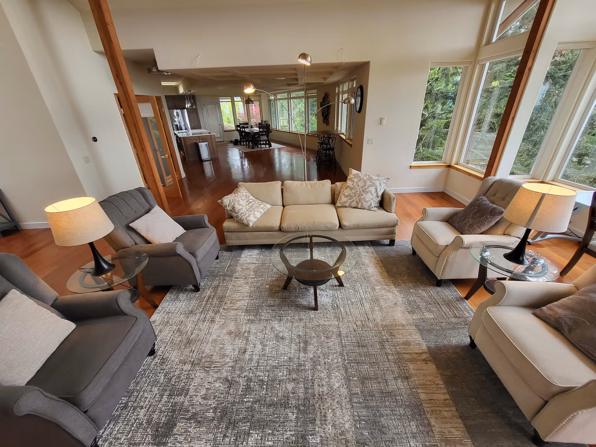 Bright open living room with a sofa, several armchairs, glass coffee tables, lamps, and large windows overlooking trees.