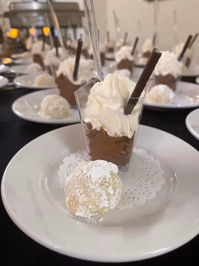 A close-up view of a dessert plate featuring a small cup of chocolate mousse topped with whipped cream and a chocolate stick, accompanied by a powdered sugar-covered cookie on a white plate with a decorative paper doily. Multiple similar dessert plates are visible in the background on a black table.