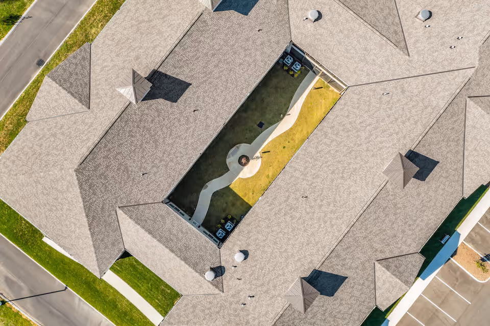 Aerial view of a senior living facility building with a central courtyard featuring a winding pathway and a circular seating area surrounded by grass. The building has a light-colored roof and is bordered by roads and parking spaces.