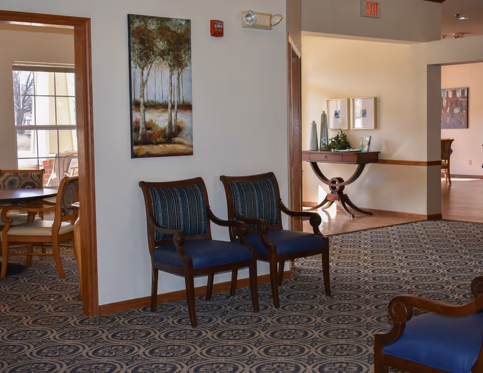 Interior view of a senior living facility showing a hallway with patterned carpet, two wooden chairs with blue cushions against a wall, a painting of trees on the wall, and a wooden table with decorative items and framed pictures in the background. There is an exit sign above a doorway leading to another room with more chairs and tables.
