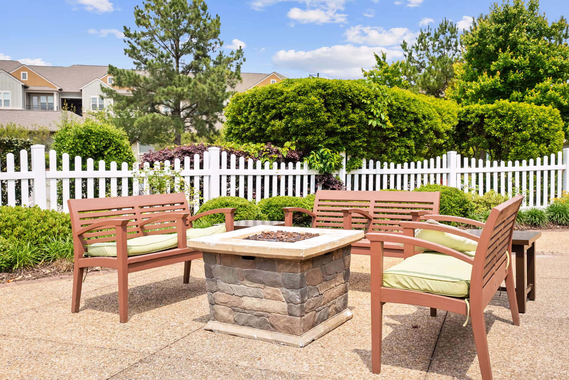 Outdoor seating area with four wooden benches with green cushions arranged around a square stone fire pit. The area is paved and surrounded by a white picket fence, green bushes, trees, and residential buildings in the background under a blue sky with scattered clouds.