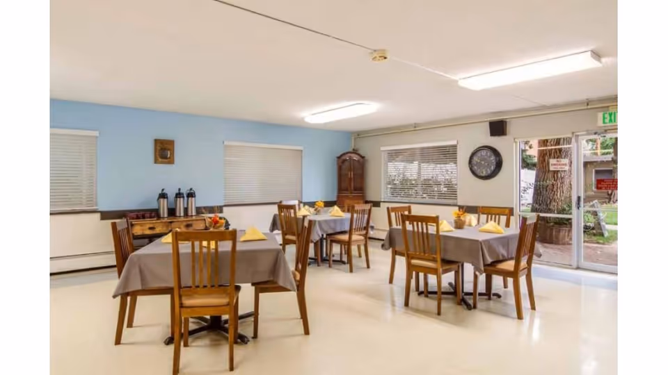 A dining room with four wooden tables covered with gray tablecloths, each set with yellow folded napkins and small flower arrangements. Wooden chairs surround the tables. The room has light blue and white walls, windows with blinds, a clock on the wall, and a glass door leading outside.
