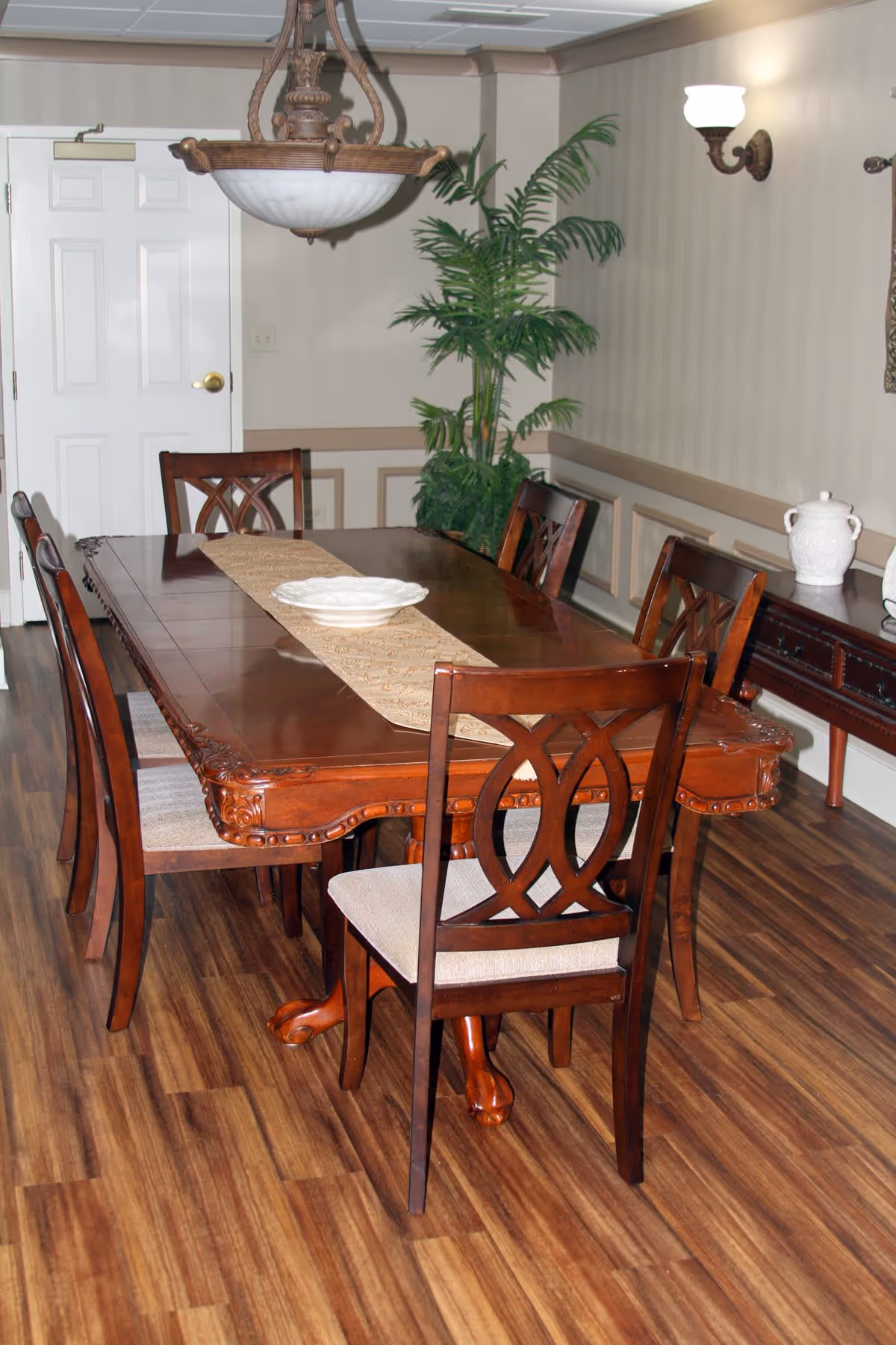A dining room with a polished wooden table and six matching chairs with cushioned seats. The table has a decorative runner and a white bowl in the center. The room features wood flooring, beige walls with decorative molding, a large potted plant in the corner, a sideboard with white ceramic pitchers, and a hanging light fixture above the table.