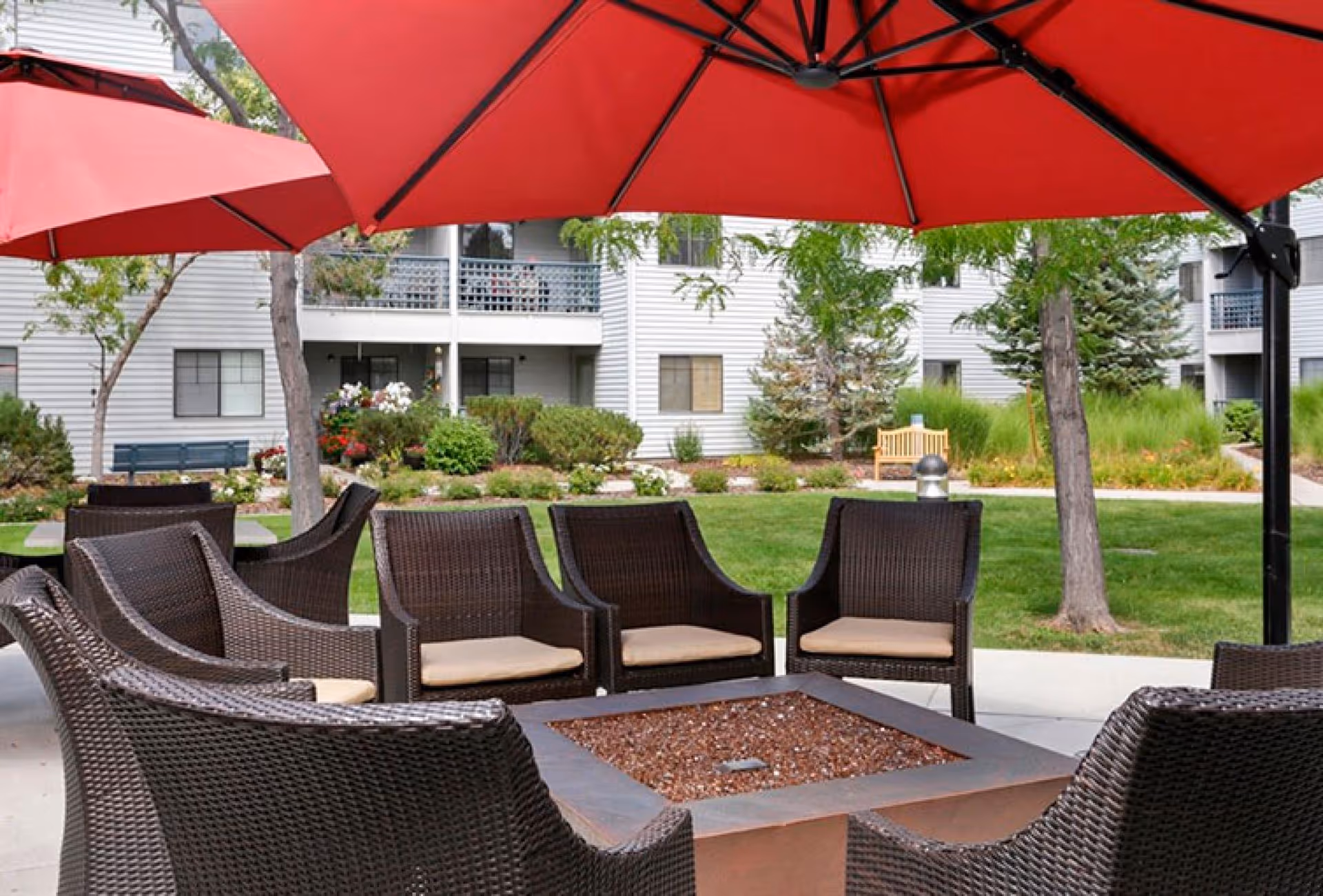 Outdoor seating area with dark wicker chairs arranged around a square fire pit table, shaded by large red umbrellas. In the background, there is a well-maintained garden with green grass, bushes, trees, and a wooden bench, with a multi-story residential building visible behind.