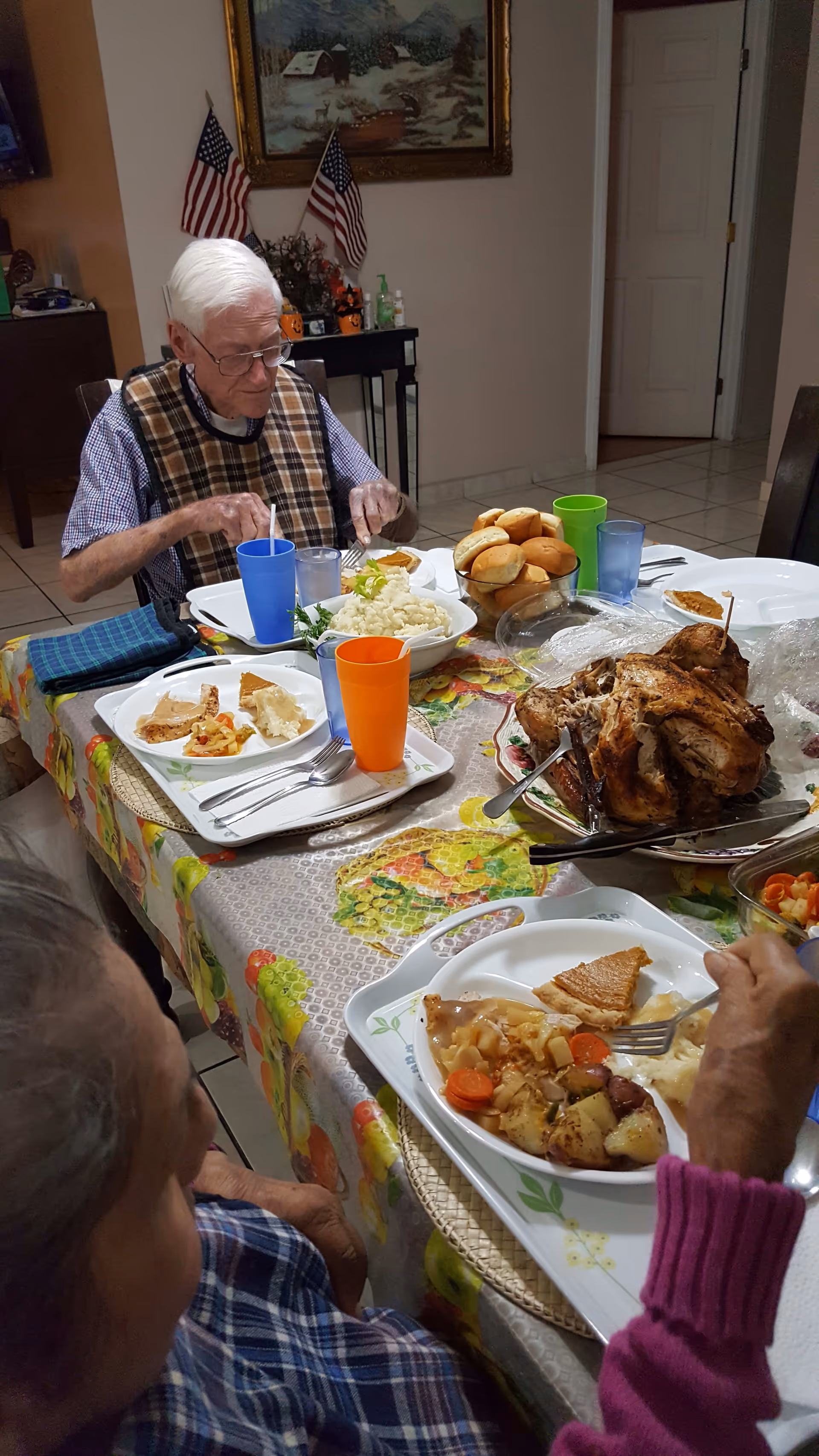 Two people seated at a dining table set with a roast chicken, rolls, mashed potatoes, vegetables and colorful cups in a homey dining room.