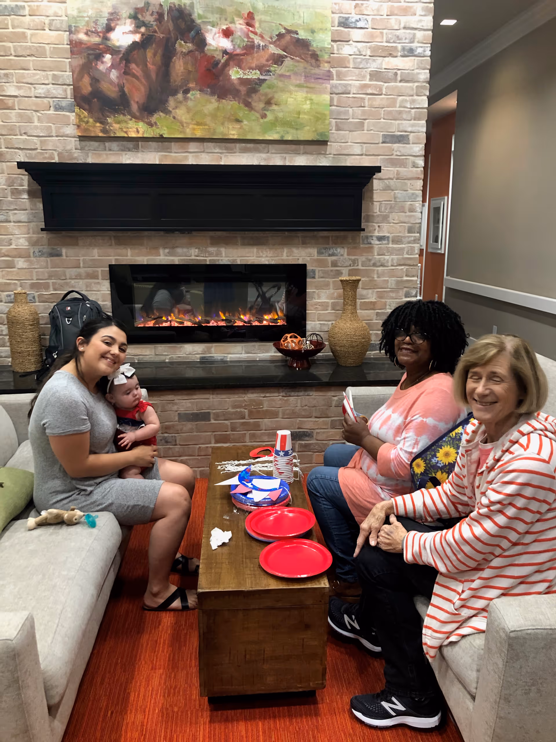Three women and a baby sit around a coffee table in a cozy lounge area with a brick fireplace and artwork.
