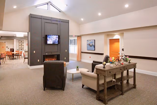 A spacious senior living common area with a high vaulted ceiling and recessed lighting. The room features a dark gray fireplace wall with a mounted TV above it. In front of the fireplace are two armchairs and a small round coffee table. To the right, there is a wooden console table decorated with flowers and a small globe. In the background, a dining area with tables and chairs is visible.