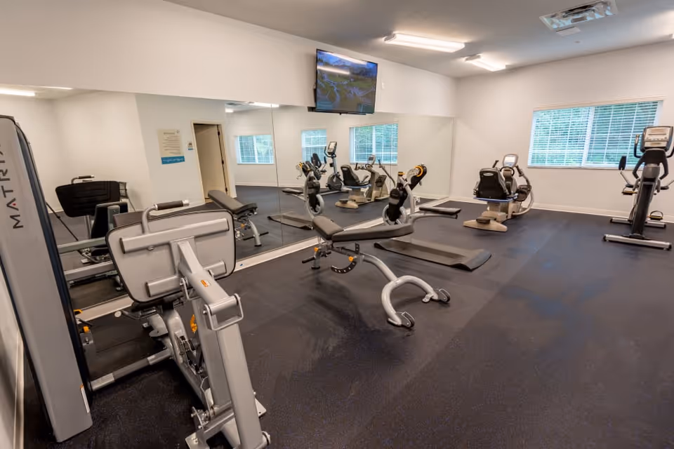 A fitness room with various exercise equipment including stationary bikes, a weight bench, and other workout machines. The room has large mirrors on one wall, a window with blinds, and a wall-mounted TV.