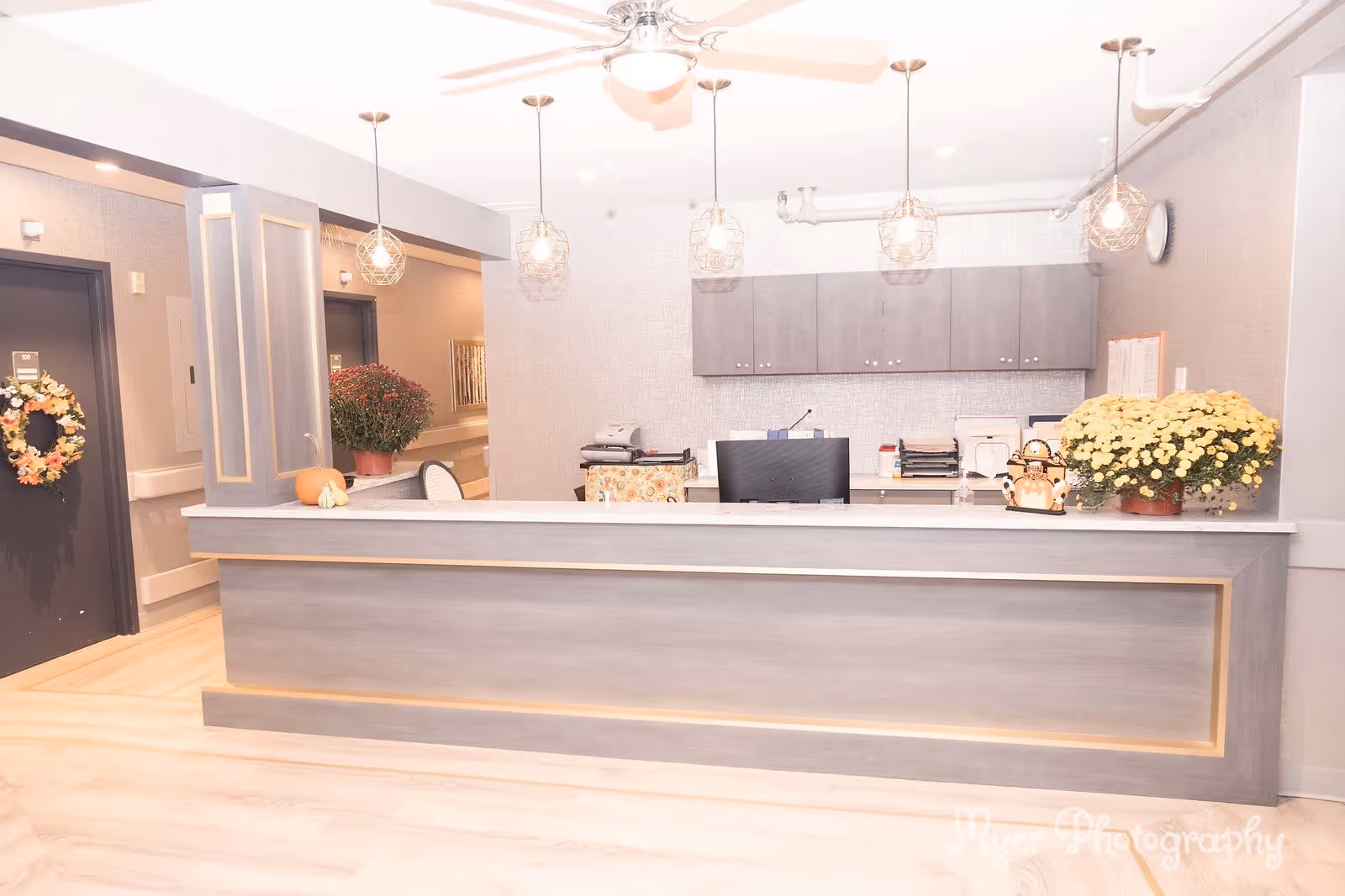 Reception desk area in a senior living facility with a gray counter, hanging pendant lights, a ceiling fan, and cabinets on the wall behind. There are decorative plants, a pumpkin, and a small Halloween-themed decoration on the counter. Doors and a hallway are visible in the background.