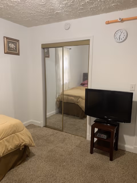 A bedroom with beige carpet and white walls. There is a bed with a beige comforter on the left side. A mirrored sliding closet door is in the center of the image, reflecting the bed and a window with sheer curtains. On the right side, a small wooden stand holds a flat-screen TV and a digital clock. A round wall clock is mounted above the TV on the wall.