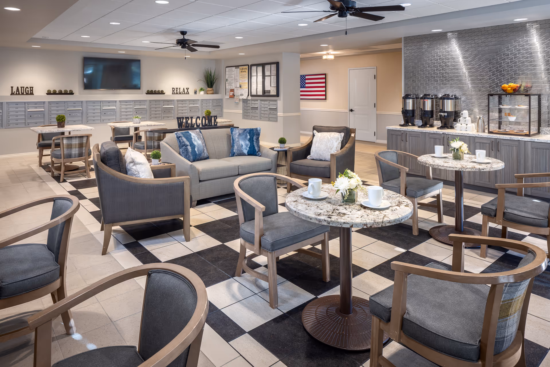 A spacious senior living common area with multiple seating options including armchairs and sofas around small round tables with white cups and flower arrangements. The floor has a checkered tile pattern. In the background, there are mailboxes on the wall with decorative words like 'LAUGH', 'RELAX', and 'WELCOME'. A beverage station with coffee dispensers and cups is visible on the right side. The room is well-lit with ceiling lights and a ceiling fan.