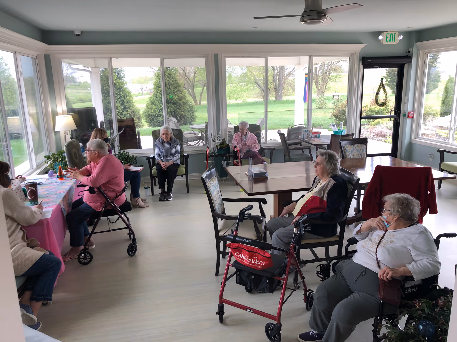 Several elderly residents seated and socializing in a bright assisted-living common room with large windows overlooking green grounds.