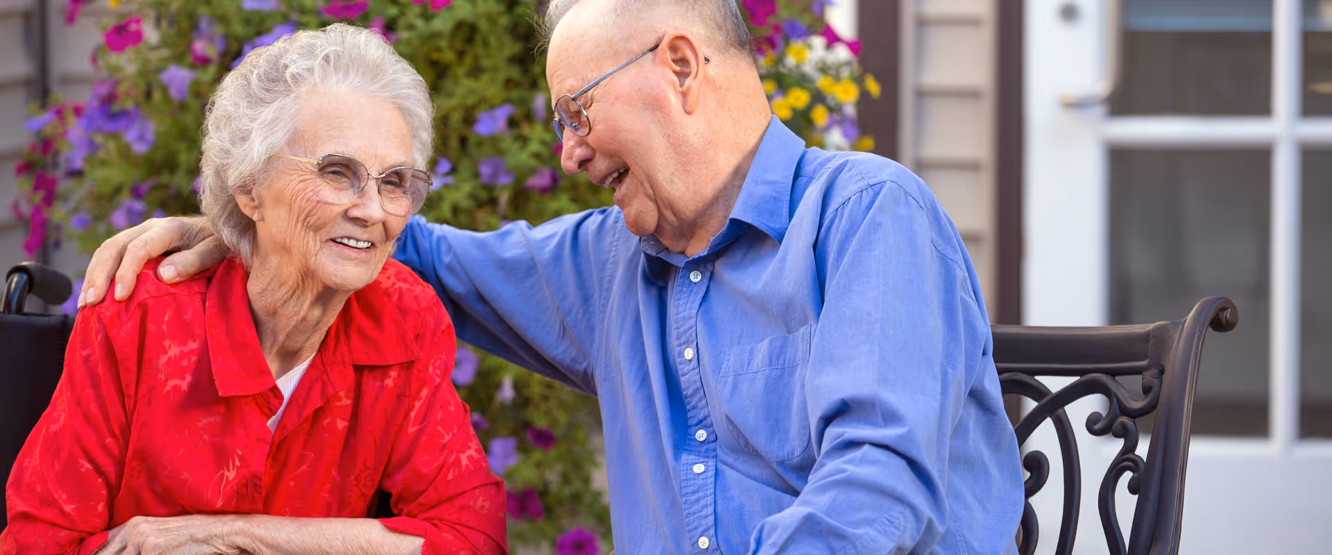 An elderly man and woman sitting outside on a bench, smiling and enjoying each other's company. The man is wearing a blue shirt and glasses, with his arm around the woman who is wearing a red blouse and glasses. Behind them are green plants and purple flowers, with a building door visible in the background.