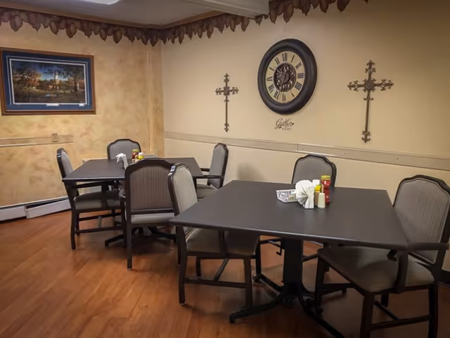 A dining area with two dark wood tables, each surrounded by four cushioned chairs. The tables have condiments and napkins on them. The walls are decorated with a large clock, two ornamental crosses, and a framed landscape painting. The floor is wooden, and the room has a warm, cozy atmosphere.