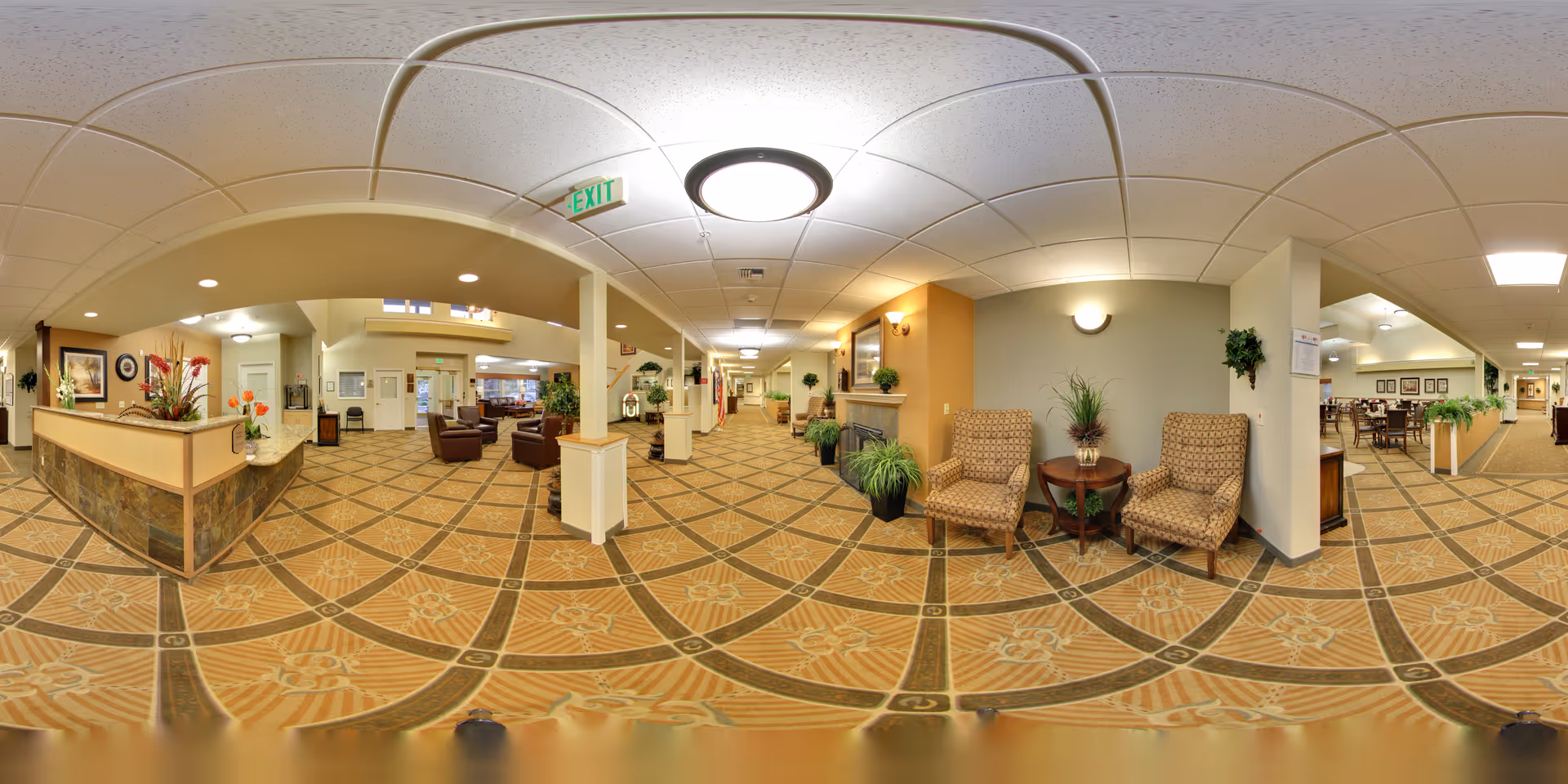 Panoramic view of a senior living facility interior showing a spacious lobby area with a reception desk on the left, comfortable seating areas with armchairs and small tables, decorative plants, and a hallway leading to other rooms. The floor is carpeted with a patterned design, and the ceiling has recessed lighting and a central light fixture. There is an exit sign above a doorway in the middle of the image.
