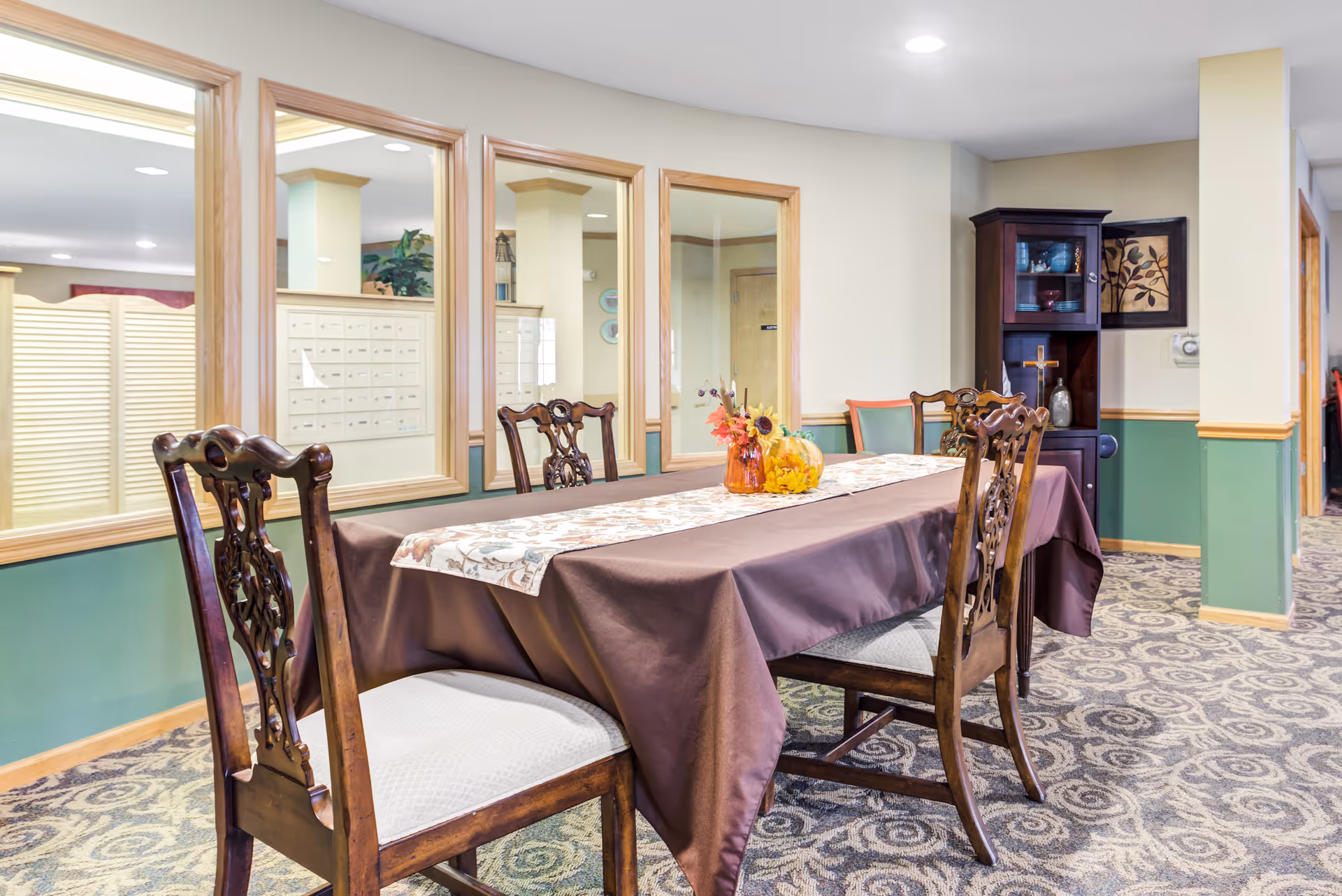 A dining area in a senior living facility with a rectangular table covered by a brown tablecloth and a decorative runner. Four wooden chairs with cushioned seats surround the table. The table is decorated with a small autumn-themed centerpiece including sunflowers and a pumpkin. The room has green and beige walls with wooden trim, patterned carpet, and large windows looking into another room with mailboxes. A dark wooden cabinet with decorative items and a framed artwork is visible in the background.