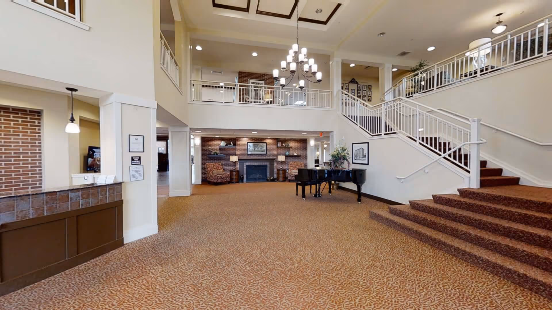 Spacious senior living facility lobby with a grand piano, carpeted floor, staircase with white railings, two armchairs by a brick fireplace, and a large chandelier hanging from a high ceiling.
