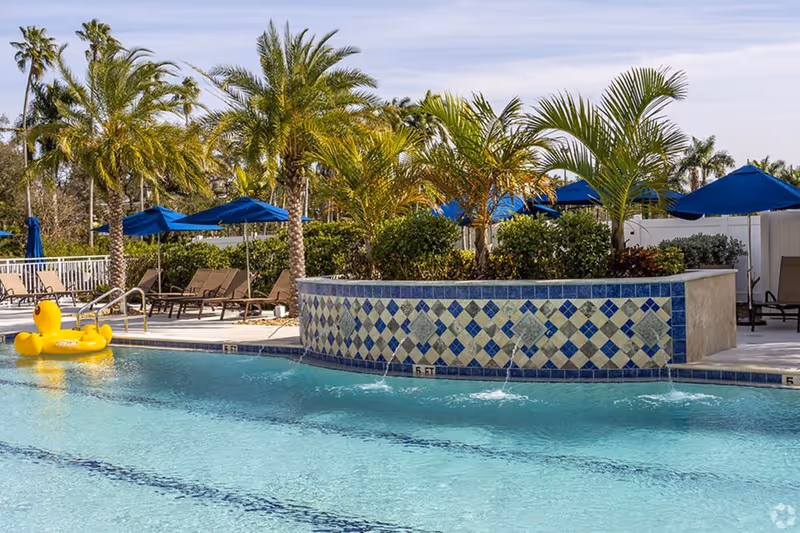 Outdoor swimming pool with clear blue water, a yellow inflatable duck float, surrounded by palm trees, blue umbrellas, and lounge chairs under a partly cloudy sky.