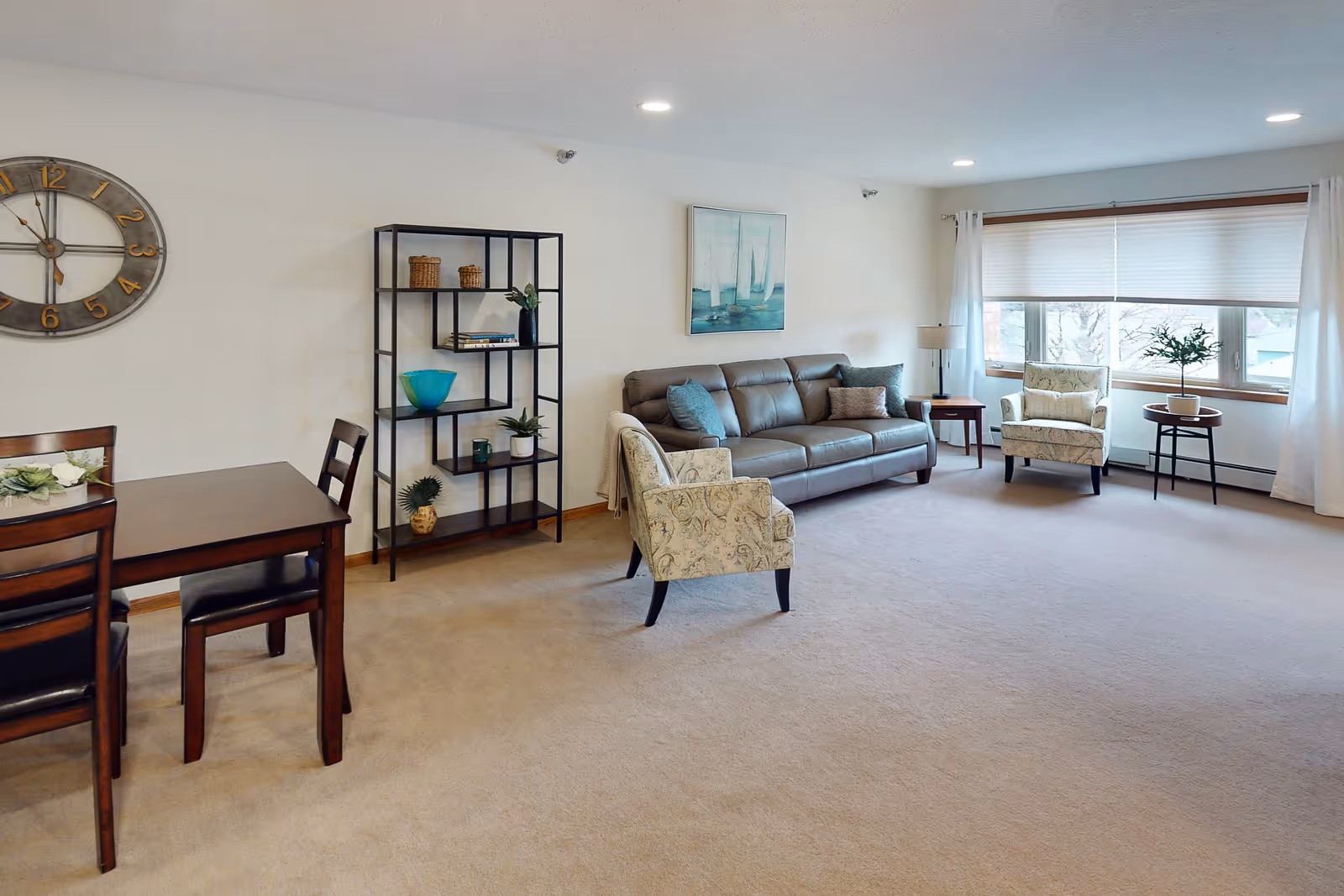 A spacious living room with beige carpet, a brown leather sofa with blue and beige cushions, two patterned armchairs, a black metal shelving unit with decorative items, a wooden dining table with chairs, a large wall clock, a painting of sailboats, and a large window with white curtains letting in natural light.