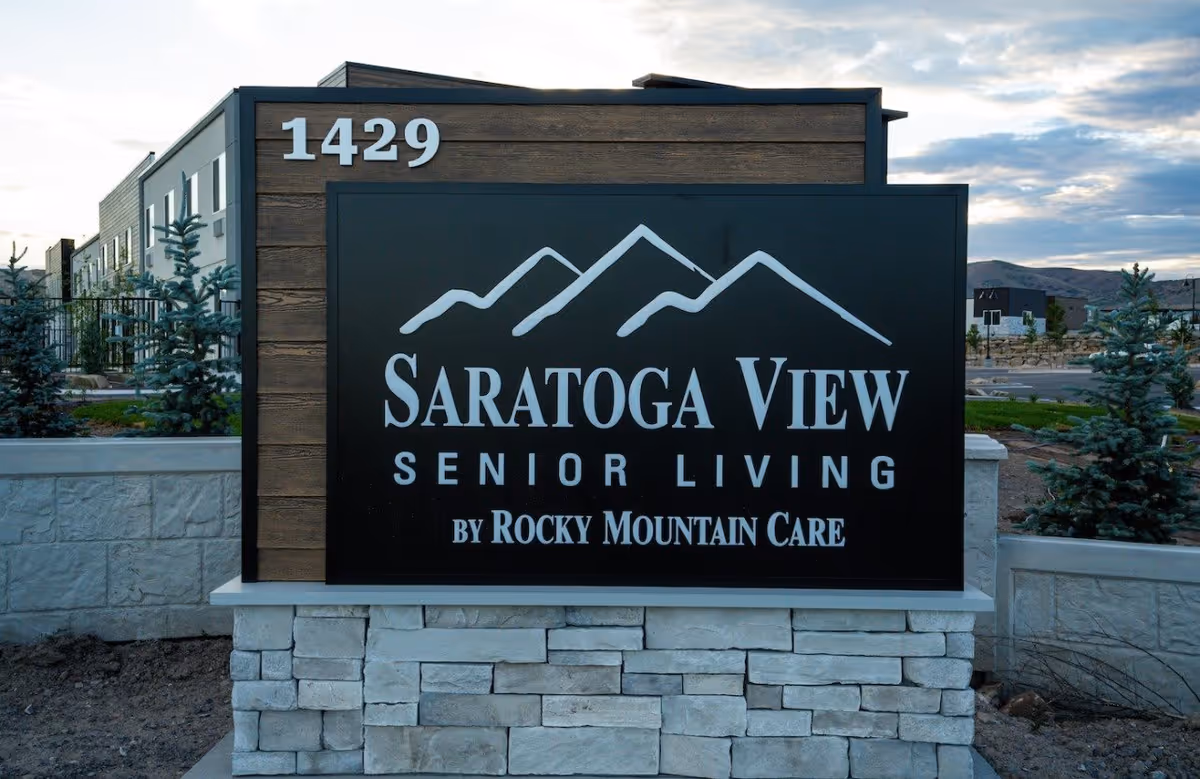 Outdoor sign for Saratoga View Senior Living by Rocky Mountain Care, mounted on a stone base with a wooden backdrop, with some small pine trees and a building visible in the background under a partly cloudy sky.