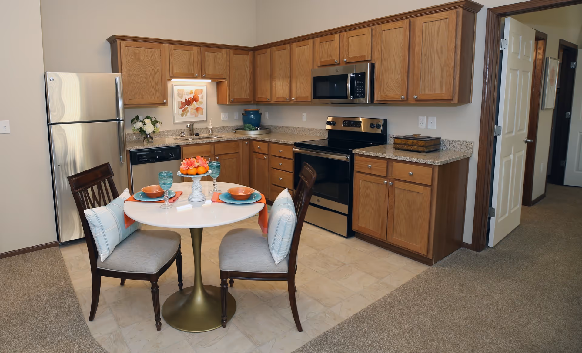 A kitchen area in a retirement community featuring wooden cabinets, a stainless steel refrigerator, stove, microwave, and dishwasher. There is a small round dining table set for two with plates, glasses, and a centerpiece of flowers and fruit. The floor is a combination of tile in the kitchen area and carpet in the adjacent hallway.