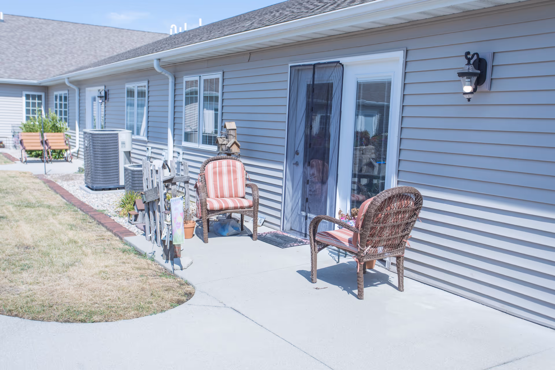 Outdoor patio area of a senior living facility with two wicker chairs with red and white striped cushions placed on a concrete walkway next to a building with gray siding. There are decorative items and potted plants near the chairs, a glass door with a mesh screen, and a wall-mounted light fixture. In the background, there are additional benches and air conditioning units along the building.