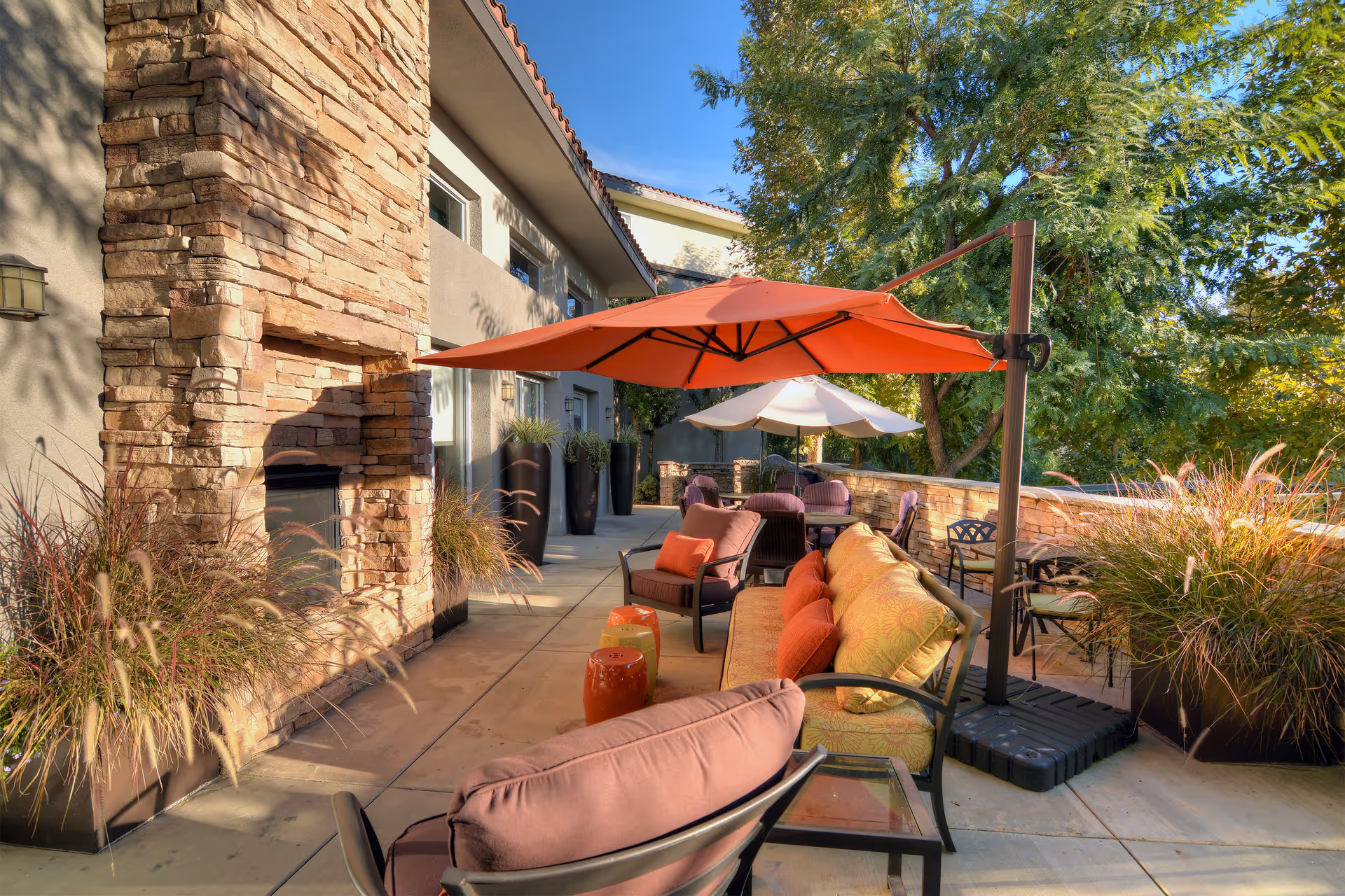 Outdoor patio area with cushioned seating, orange and white umbrellas, a stone fireplace, potted plants, and trees in the background under a clear blue sky.