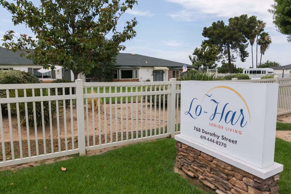 Outdoor view of Lo-Har Senior Living facility showing a white fence, green lawn, trees, and a sign with the facility's name, address, and phone number.