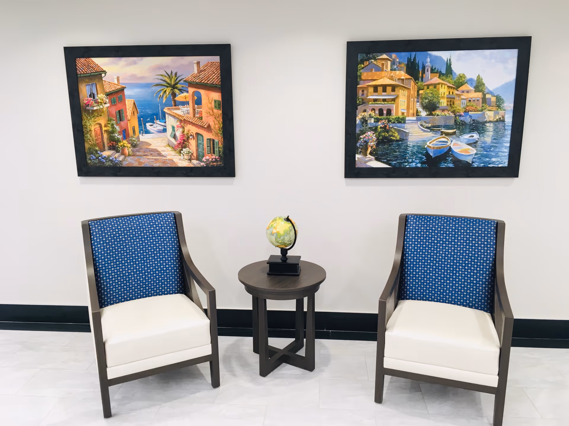 Two wooden armchairs with blue patterned backs and white seats are placed on either side of a small round wooden table with a decorative globe on top. Behind the chairs, two framed paintings of colorful coastal village scenes hang on a plain white wall.
