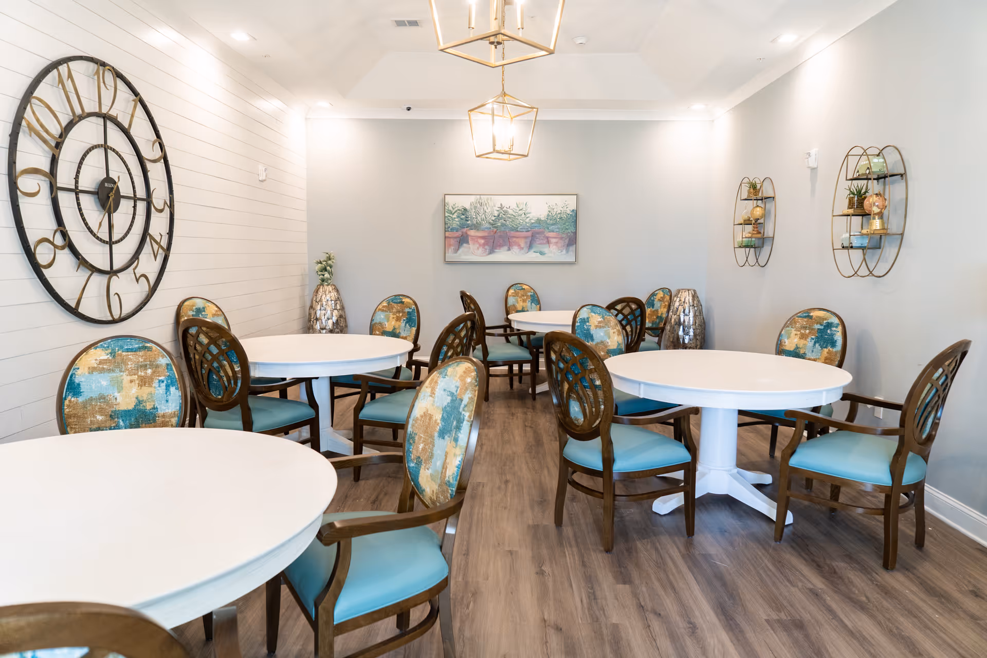 A bright dining room with round white tables and wooden chairs featuring blue and gold patterned cushions. The room has light gray walls, wood flooring, a large decorative wall clock, two modern pendant lights, wall shelves with small plants and decor, and a framed painting of potted plants on the far wall.