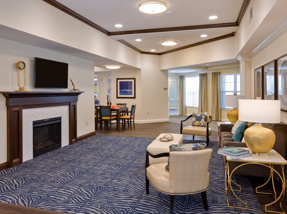 A spacious living room area with a patterned blue carpet, beige armchairs with decorative pillows, a brown sofa, and a gold table lamp on a side table. There is a fireplace with a mounted flat-screen TV above it, and a dining table with chairs is visible in the background. The room has large windows with beige curtains and recessed ceiling lights.