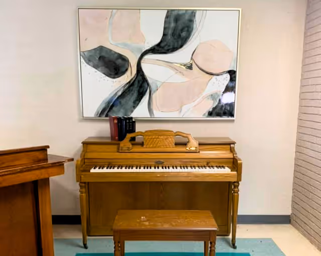 Upright wooden piano with a bench below and an abstract painting hanging on the wall above in a room.