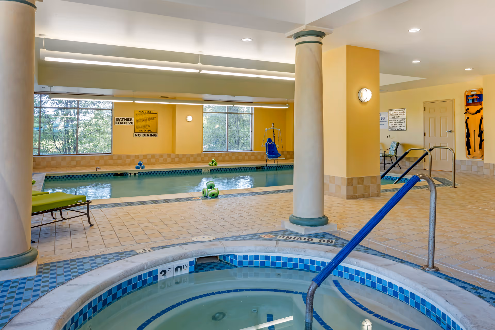 Indoor pool area with a small hot tub in the foreground, a swimming pool in the background, yellow walls, tiled floors, large windows, pool safety signs, and pool equipment including a pool lift chair.