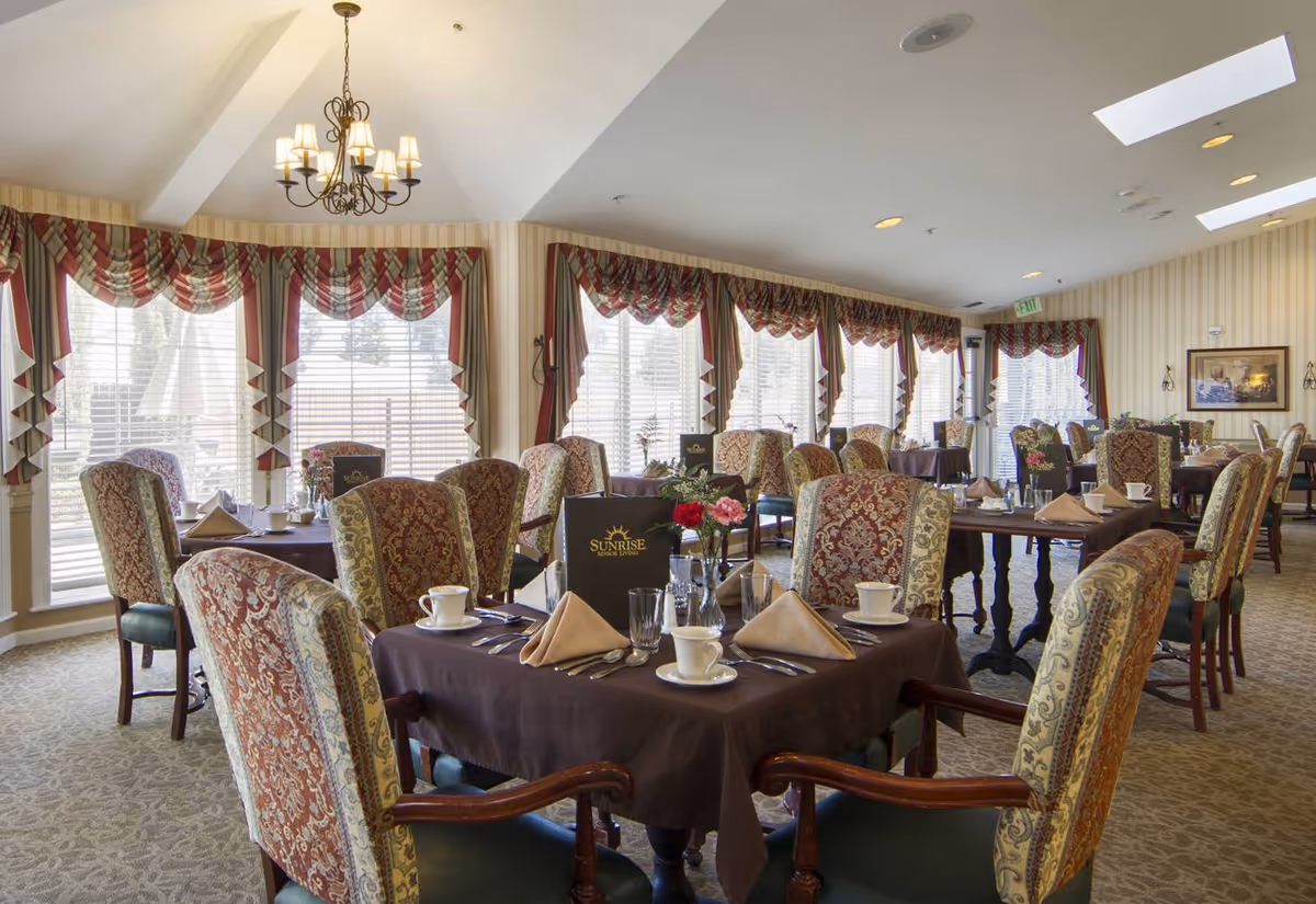A well-lit dining room with multiple tables covered in dark tablecloths, each set with beige napkins, white cups, glasses, and silverware. The chairs have ornate upholstery with red and gold patterns. Large windows with red and beige draped curtains allow natural light to fill the room. A chandelier hangs from the ceiling, and menus labeled 'Sunrise' are placed on the tables.