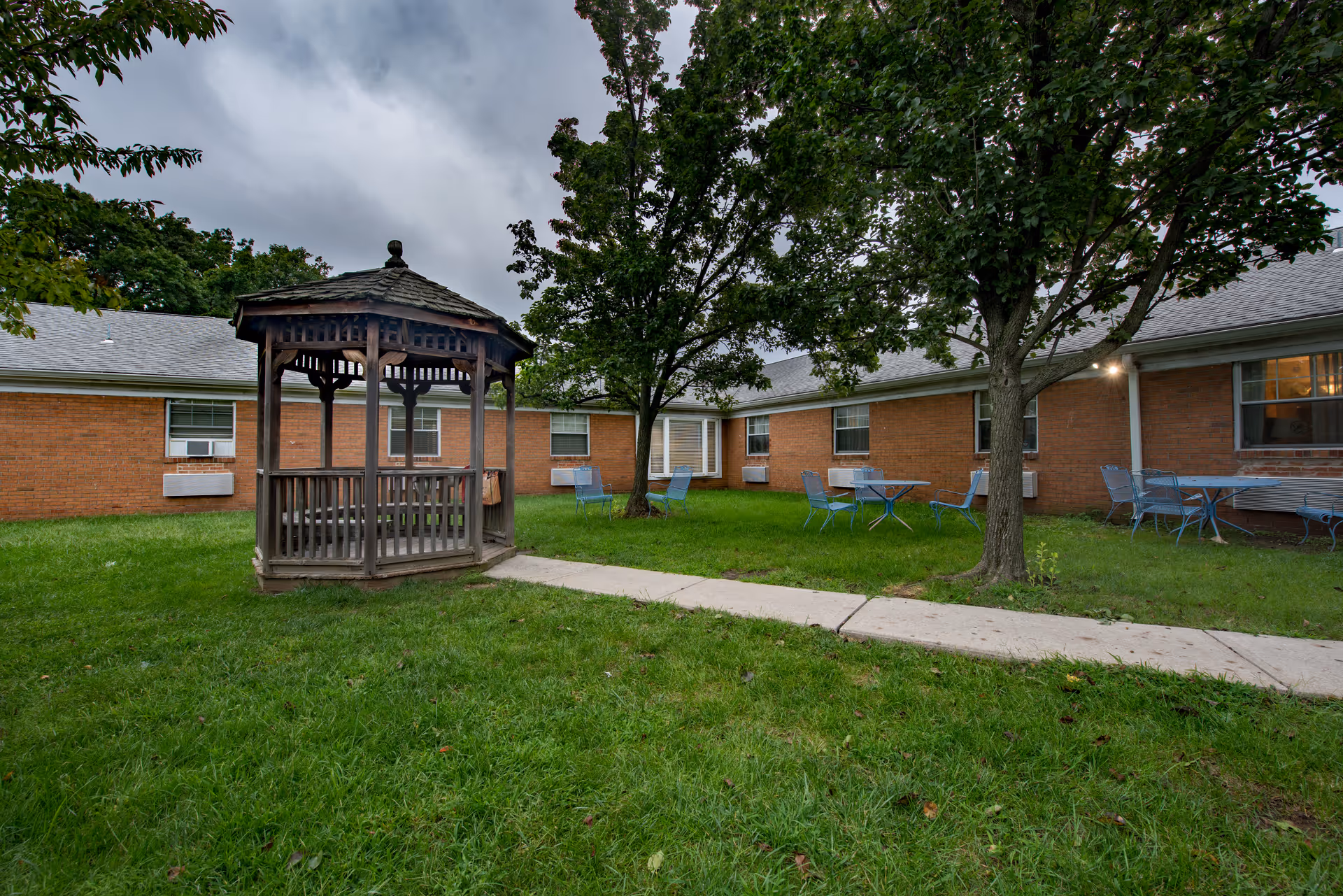 Outdoor courtyard area with a wooden gazebo, green grass, trees, and several blue metal tables and chairs. The courtyard is surrounded by a single-story brick building with multiple windows and air conditioning units.