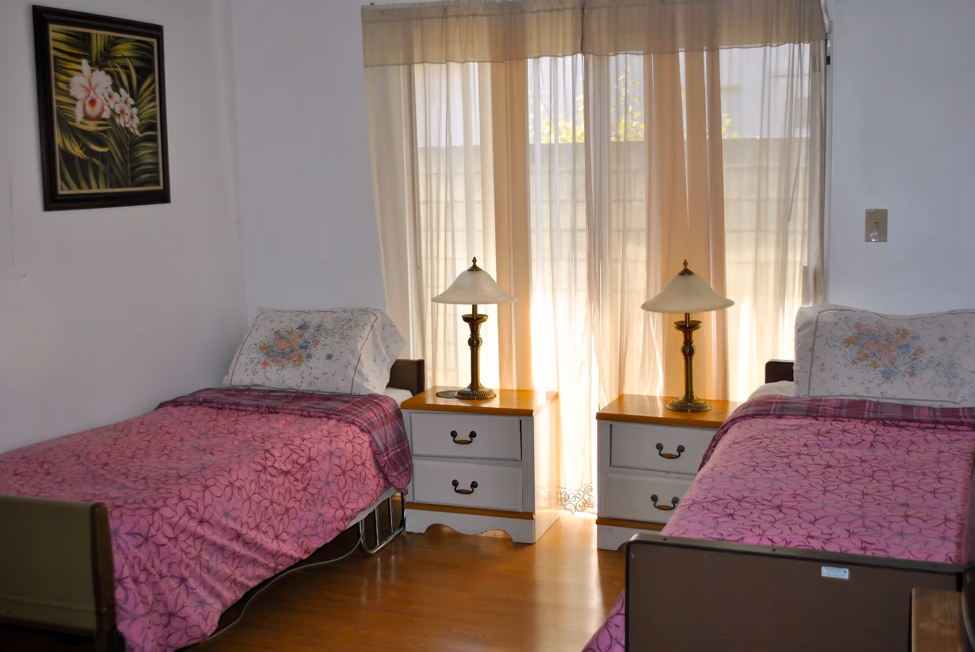 A bedroom with two single beds, each covered with a pink floral bedspread and a floral pillow. Between the beds are two white nightstands with wooden tops, each holding a brass lamp with a white lampshade. A large window with sheer beige curtains is behind the nightstands, allowing natural light to enter the room. A framed painting of flowers and leaves hangs on the left wall.