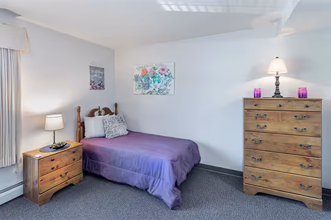 A simple bedroom with a single wooden bed covered with a purple bedspread and a decorative pillow. There is a wooden nightstand with a lamp and a small plant on it to the left of the bed. On the right side of the room, there is a tall wooden dresser with a lamp and two purple candles on top. The walls are white with two pieces of floral artwork hanging above the bed. The room has a gray carpeted floor and a window with light-colored curtains.