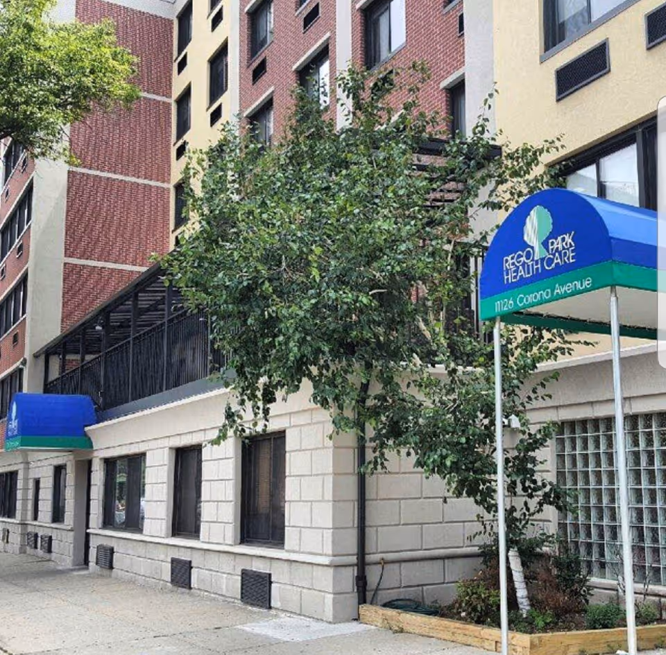 Exterior view of a multi-story building with a blue and green awning that reads 'Rego Park Health Care 11126 Corona Avenue'. There is a tree and some landscaping in front of the building.