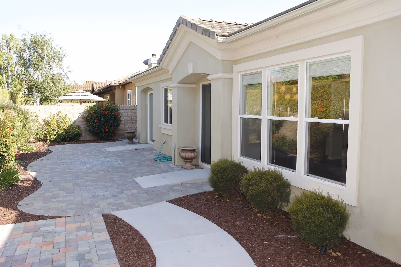 Outdoor patio area of a senior living facility with a paved walkway, beige building exterior, windows, and landscaped bushes and plants along the path.