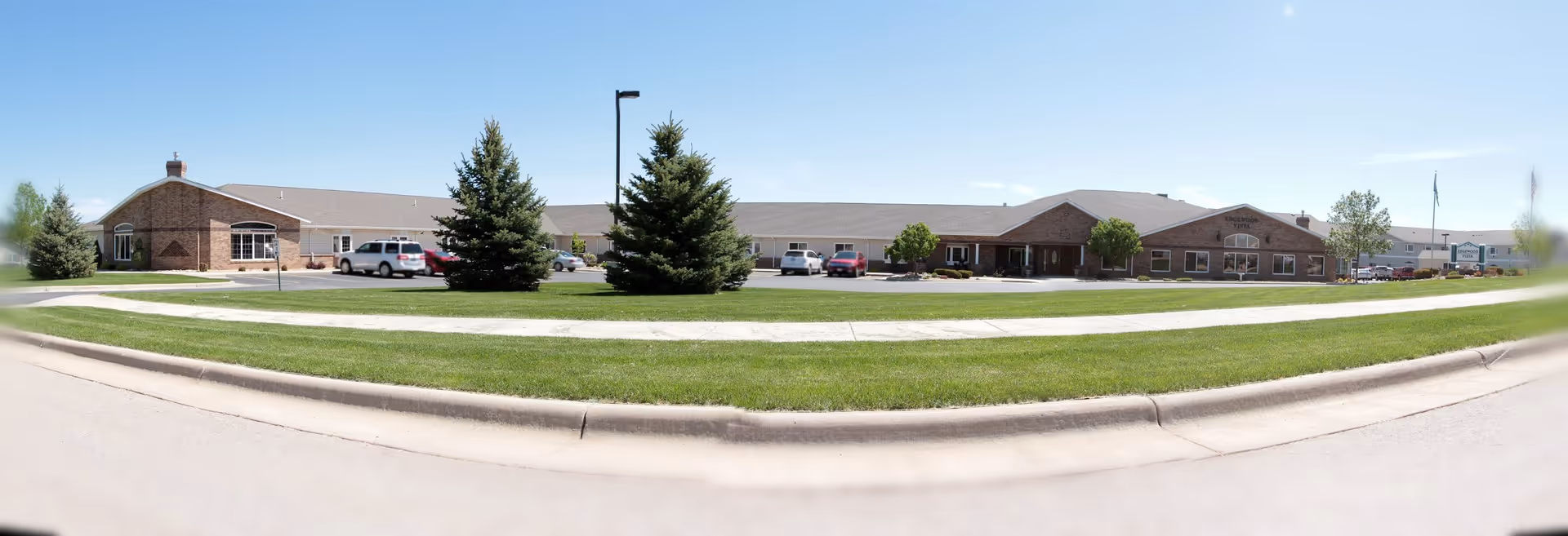 Wide exterior view of a single-story senior living facility building with brick and siding facade, surrounded by green grass, trees, and a parking lot with several cars. The sky is clear and blue.