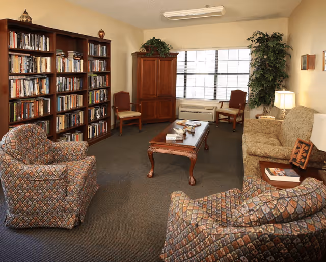 A cozy living room with patterned armchairs, a floral sofa, a wooden coffee table with books and magazines, a large bookshelf filled with books, two wooden chairs, a wooden cabinet, a large window with blinds, a potted plant, and a table lamp providing warm lighting.