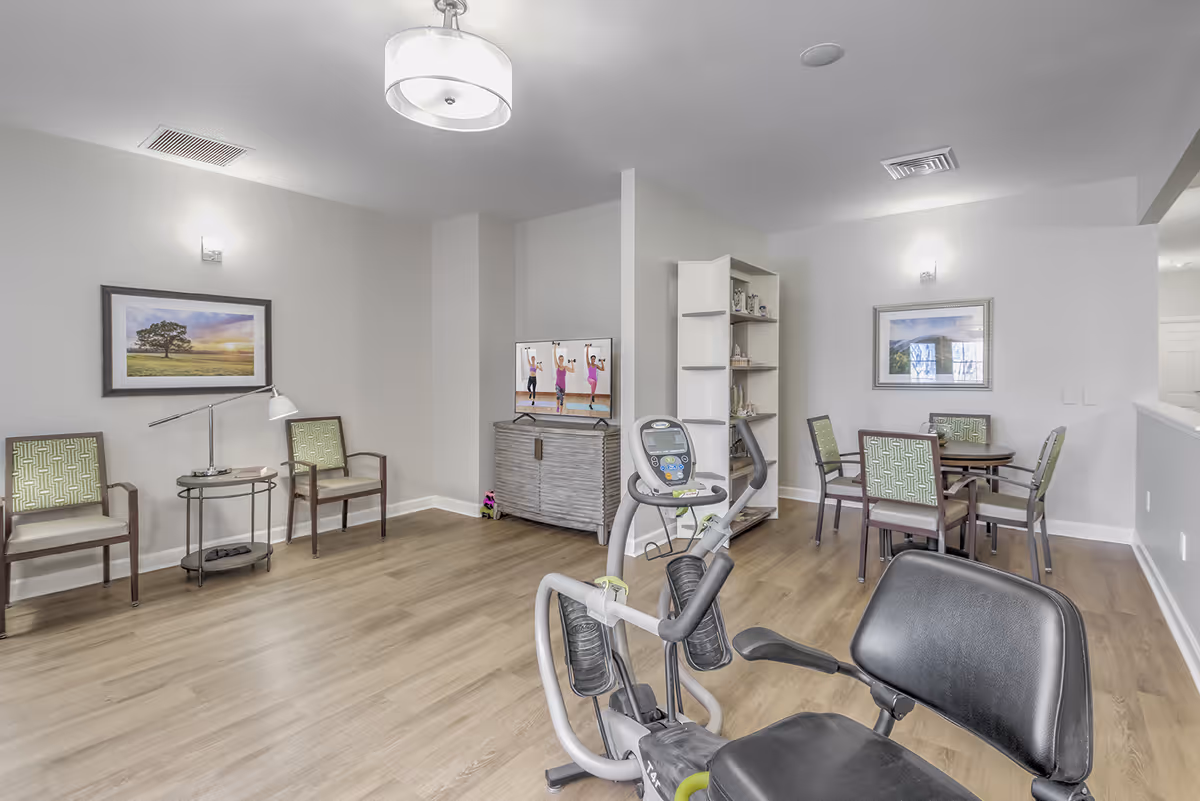 A bright and clean room with light wood flooring featuring a recumbent exercise bike in the foreground. The room has two seating areas with green patterned chairs and small tables, a TV on a cabinet displaying an exercise video, and a white shelving unit with decorative items. The walls are light gray with framed landscape artwork and modern light fixtures.