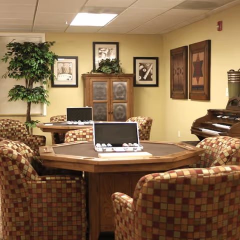 A cozy common area with a wooden octagonal game table surrounded by cushioned chairs with a checkered pattern. Two open cases with poker chips are on the table. The room has beige walls decorated with framed pictures and a wooden cabinet with plants on top. A piano is visible on the right side of the room.