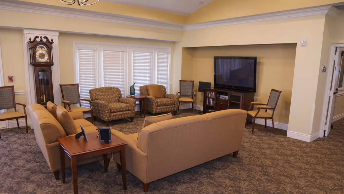A cozy living room area with beige sofas and patterned armchairs arranged around a wooden side table and a TV mounted on a wooden stand. The room features a grandfather clock, several chairs, and large windows with white blinds, all set against light yellow walls and carpeted flooring.