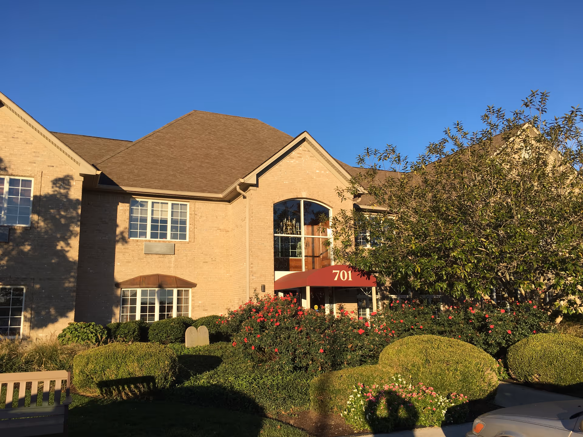 Front exterior of a brick building with a red awning reading "701", landscaped bushes and trees under a clear blue sky.