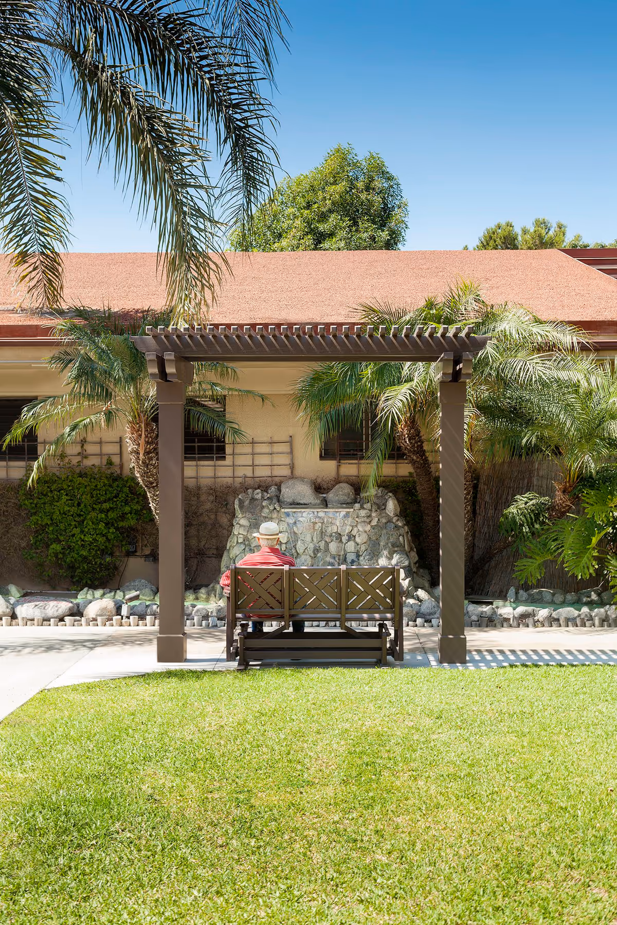 An elderly person wearing a hat and red shirt sits on a bench under a wooden pergola in a garden area with green grass, palm trees, and a stone water feature in the background.