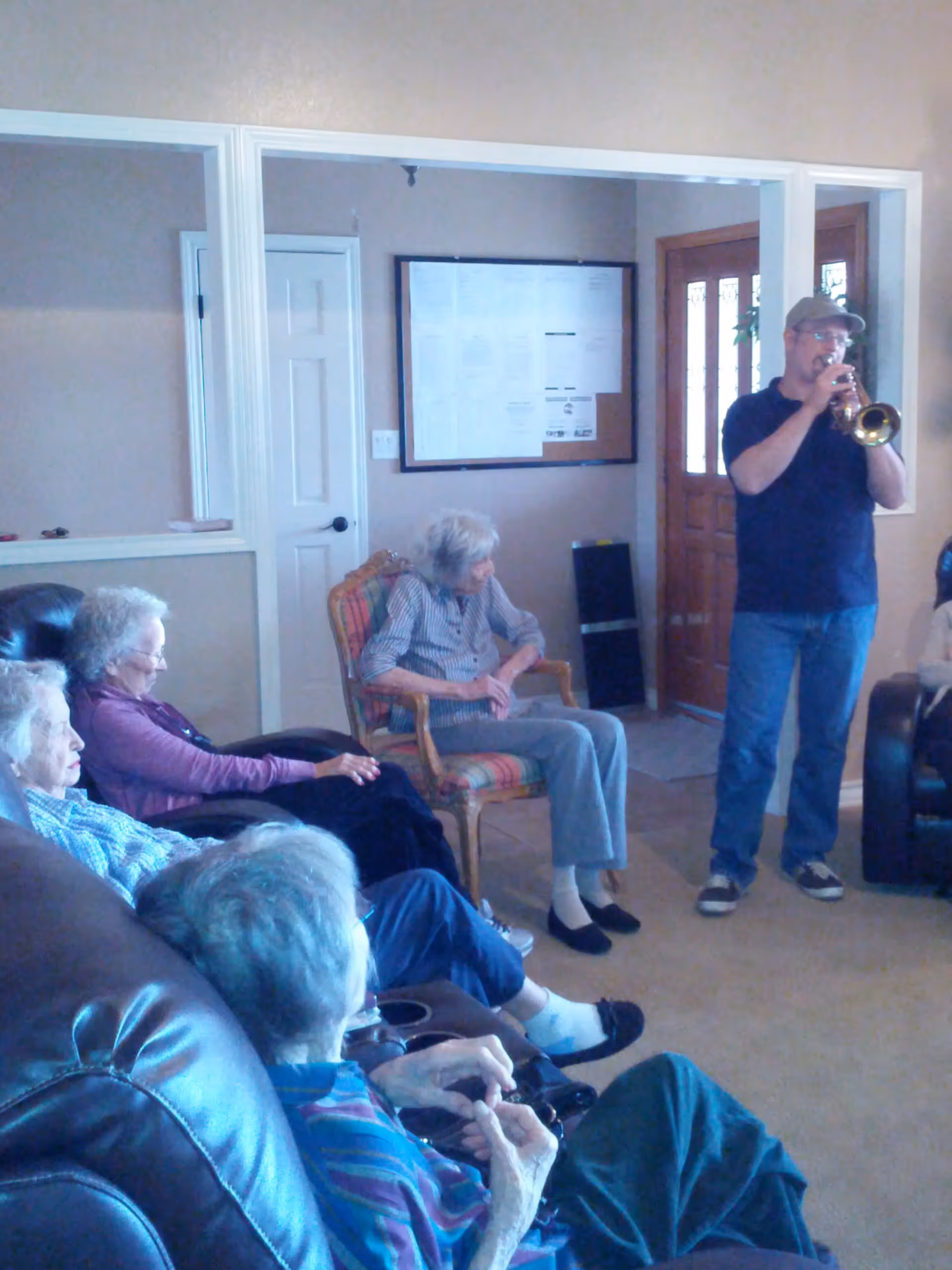 A group of elderly people sitting in a living room area, attentively watching a man playing a trumpet near the entrance door.
